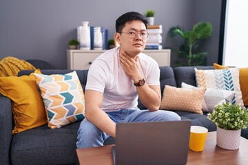 Young asian man using laptop at home sitting on the sofa touching painful neck, sore throat for flu, clod and infection