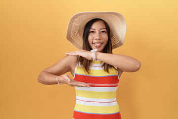 Middle age chinese woman wearing summer hat over yellow background gesturing with hands showing big and large size sign, measure symbol. smiling looking at the camera. measuring concept.