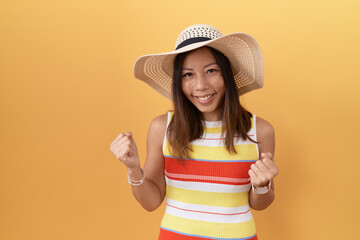 Middle age chinese woman wearing summer hat over yellow background very happy and excited doing winner gesture with arms raised, smiling and screaming for success. celebration concept.