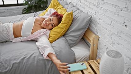 Vibrant young redhead woman lounging in bed, joyfully texting away on her smartphone in the heart of a cosy bedroom, radiating fun and confidence.
