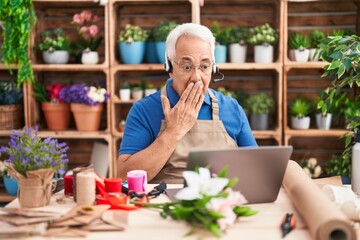 Middle age man with grey hair working at florist shop doing video call covering mouth with hand,...