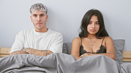 A tattooed man and a woman in lingerie sit in a bed looking upset, implying a couple in a bedroom...