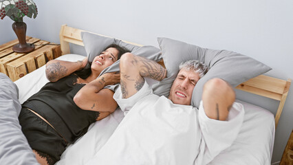 A distressed man and woman cover their ears with pillows in a bedroom, indicating annoyance or...