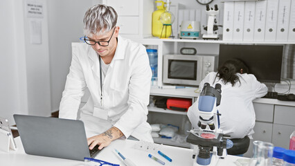 Fototapeta na wymiar A man and woman working as coworkers in a laboratory, with him using a laptop and her examining samples under a microscope.