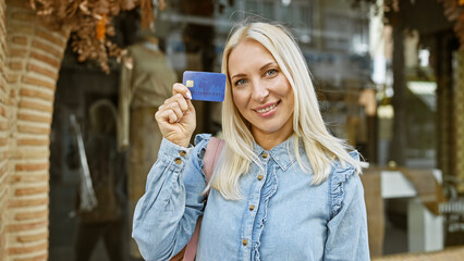Confident young blonde woman joyfully holding a credit card, smiling radiantly in the sunlit city...