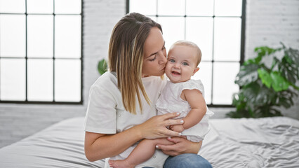 Joyful mother hugging and kissing daughter, enjoying a casual and fun moment sitting on bed in cozy bedroom, expressing confident love and positive family relationship.