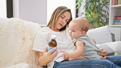 Cool mom sitting with lovely daughter on sofa, reading syrup bottle label, chatting away on phone in cozy home