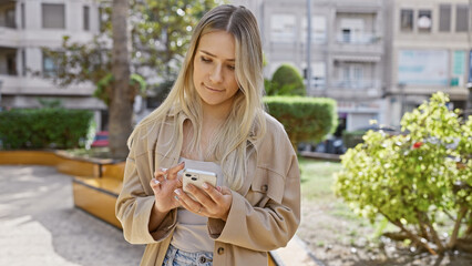 Beautiful blonde, a young woman with a cool, casual lifestyle, standing seriously in a sunny park, fully concentrated on her smartphone while texting online