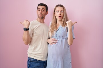 Young couple expecting a baby standing over pink background surprised pointing with hand finger to...