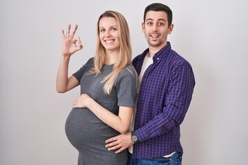 Young couple expecting a baby standing over white background smiling positive doing ok sign with hand and fingers. successful expression.