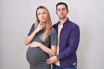 Young couple expecting a baby standing over white background thinking concentrated about doubt with finger on chin and looking up wondering