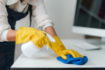 Close-up of hands in yellow gloves cleaning a white desk with a spray bottle and blue cloth, emphasizing cleanliness and hygiene..