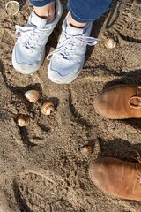 A top-down view of two different pairs of shoes on sandy beach ground with scattered seashells
