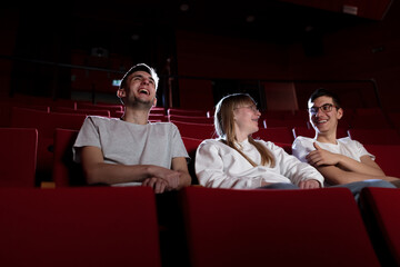 Three friends, a teenage girl and two boys smiling, watching a movie in an empty cinema with red chairs. Young adults and leisure concepts. - obrazy, fototapety, plakaty