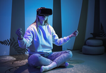 Girl wearing a virtual reality headset sitting in a lotus pose and meditating in a room with blue...