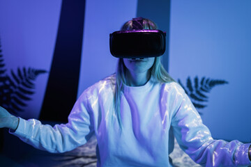 Girl wearing a virtual reality headset sitting in a lotus pose and meditating in a room with blue neon light and glowing neon lines.