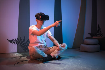 Smiling teenage programmer boy sitting on the floor with a virtual reality headset on, making hand gestures, typing, programming and scrolling in the air.