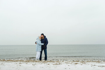 An elderly couple stands by the seashore in winter, hugging.