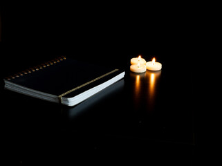 Cahier fermé sur une table noire avec 3 petites bougies chauffe plat allumées et leur reflet - fond noir et espace vide