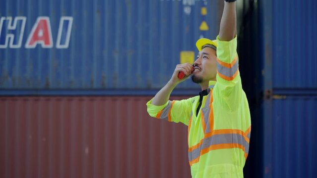 A worker wearing a hard hat and safety vest talking with a walkie-talkie is loading container boxes at a logistics warehouse on a cargo ship for import-export in the port.