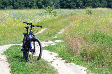 Fototapeta premium bike stands on the road in the field. A mountain bike stands on a field path with green grass. Mountain bike, blooming summer field, meadow flowers, sunny day. ride a bike. outdoor activities.