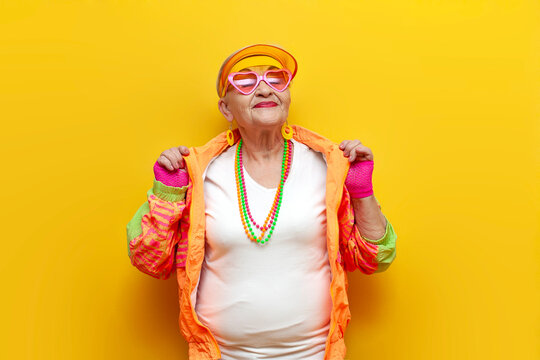 old crazy grandmother in fashionable sportswear rejoices and celebrates victory and dances on a yellow isolated background, elderly funny pensioner with glasses raises her hands up