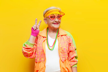 old crazy grandmother in fashionable sportswear smiles and shows peace gesture on yellow isolated background, elderly funny pensioner woman in glasses shows number two with fingers