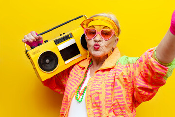 old crazy grandmother in fashionable sportswear listens loudly to music on a tape recorder and takes a selfie on yellow isolated background, elderly funny pensioner in glasses holds a boombox and