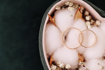 Two golden wedding rings rest on cotton balls in a heart-shaped box, surrounded by small dried...
