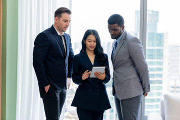 Diverse smart businesspeople standing near window with skyscraper while looking at business data analysis. Group of business team using tablet represent marketing plan, strategy, idea. Ornamented..