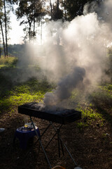 Backlit BBQ grill enveloped in smoke at sunset in a forested area, with a stool and tea glass nearby, evoking a rustic outdoor cooking scene