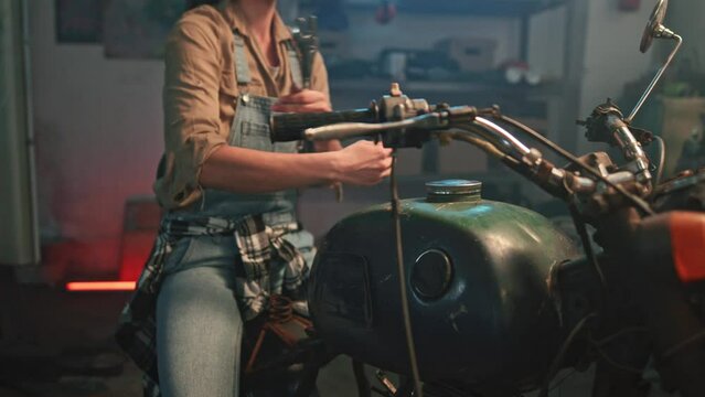 Portrait of young female auto mechanic sitting on motorcycle and holding wrench. Beautiful professional repair woman with crossed arms in garage looks directly into camera and smiles.