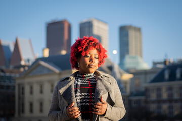 sexy moody female model with strong unique style appearance in urban inner city environment The Hague cityscape skyline. Confident black lady stylish red hair with winter good taste Holland fashion	 - obrazy, fototapety, plakaty