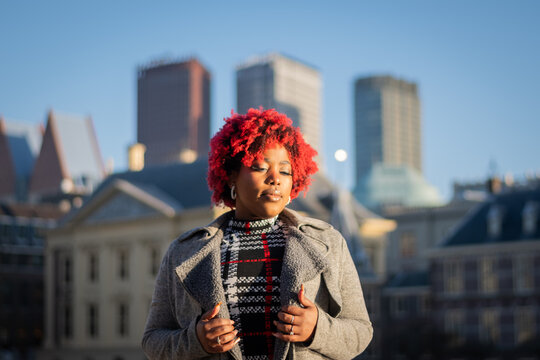sexy moody female model with strong unique style appearance in urban inner city environment The Hague cityscape skyline. Confident black lady stylish red hair with winter good taste Holland fashion	