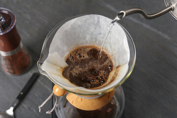 Pouring hot water into glass chemex coffeemaker with paper filter and coffee on gray table, above view