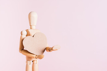 Wooden man holding craft cardboard heart in hands on pink background