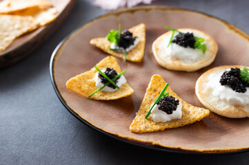 Nachos and mini pancakes with black caviar on sour cream on a plate.