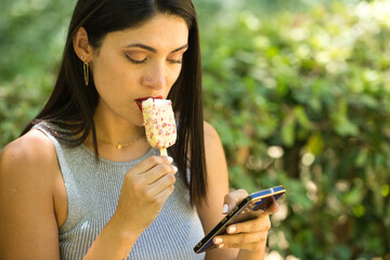 Latin woman, young and beautiful brunette consults her mobile phone and eats a refreshing ice cream to combat the high temperatures in the city. Heat wave and summer concept.