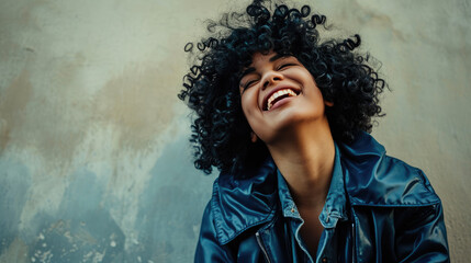 Joyful woman with curly hair is laughing and leaning against a sunlit wall. - obrazy, fototapety, plakaty