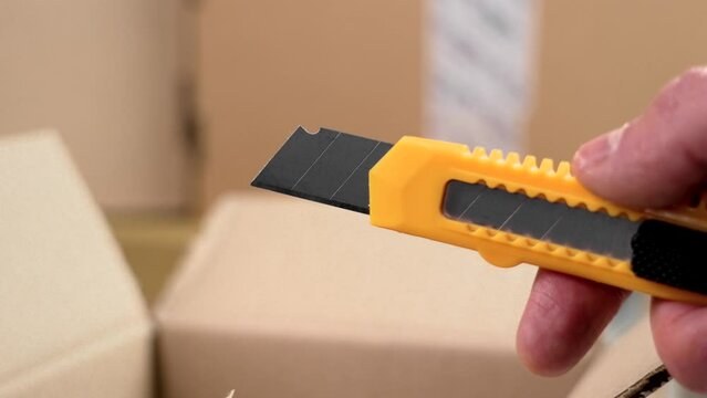 Box cutter in worker hand on a delivery warehouse on a parcel pile background close up. Retractable utility knife