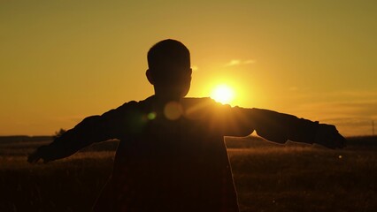 Fly concept. Child wants to become an astronaut pilot. Child boy play like an airplane, in park opposite sky at sunset. Boy aviator dreaming raised his hands to wings of plane on field in rays of sun