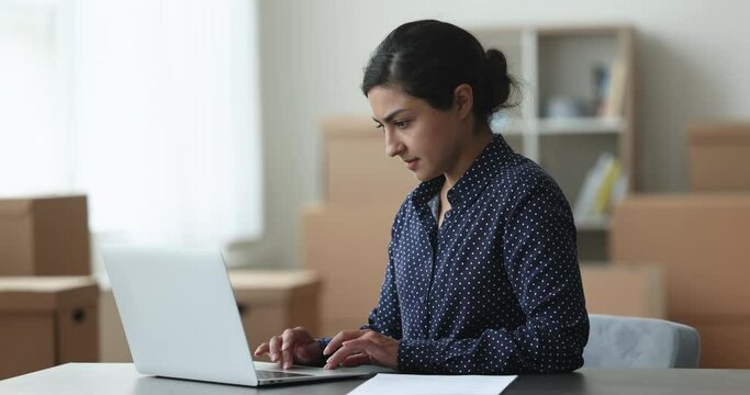 Serious young Indian businesswoman typing on laptop in e-commerce storage room with stacked paper containers behind, processing online customer order, using business application