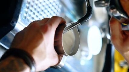 Barista's hands hold the metal pot filling it with hot milk from coffee machine. Making latte or cappuccino. Close up. Vertical view.