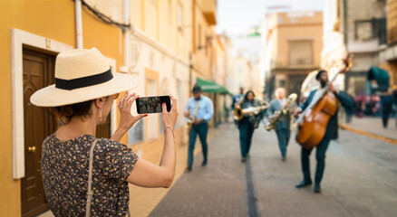 Tourist woman with straw hat taking a photograph of a group of unrecognizable musicians on the street, playing jazz. suitable for International Jazz Day on April 30 or International Music Day on