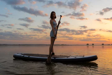 Fotobehang Ontspanning athletic woman on SUP paddle boarding through shining water at sunset. aesthetically wide shot. Freedom happy female relaxing on surfboard on a lake  © Yekatseryna