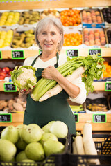 Mature lady supermarket worker standing in salesroom of fruits and vegetables department and holding vaious vegetables in hands.