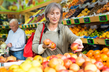 Fototapeta premium Positive female buyer shopping in supermarket, choosing ripe apples in fruit and vegetable department