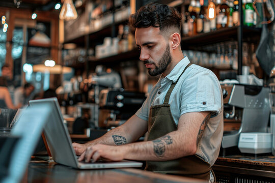 Handsome bearded barista Italian man small business owner working with laptop computer behind the counter bar in a cafe making order