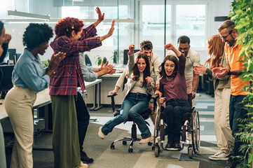 Young diverse business people racing in an office chair race at work - obrazy, fototapety, plakaty