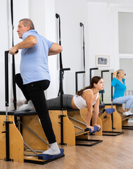 Serious concentrated elderly man wearing sportswear doing standing exercises on Pilates chair...